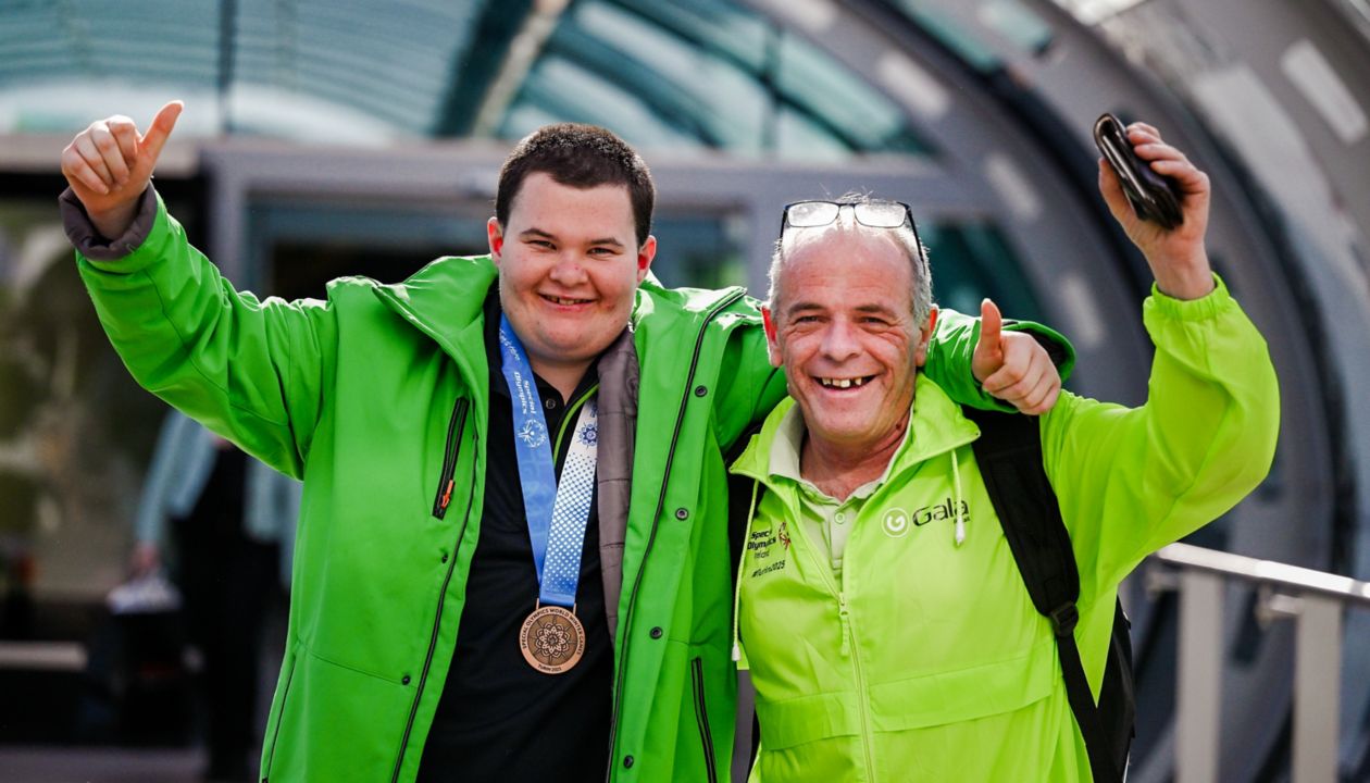 Two individuals wearing bright green jackets stand together in an outdoor setting, giving thumbs up gestures. One person displays a bronze medal on a blue and white ribbon around their neck. The background features a modern, glass-covered walkway, suggesting an event or competition. The mood is celebratory and positive.